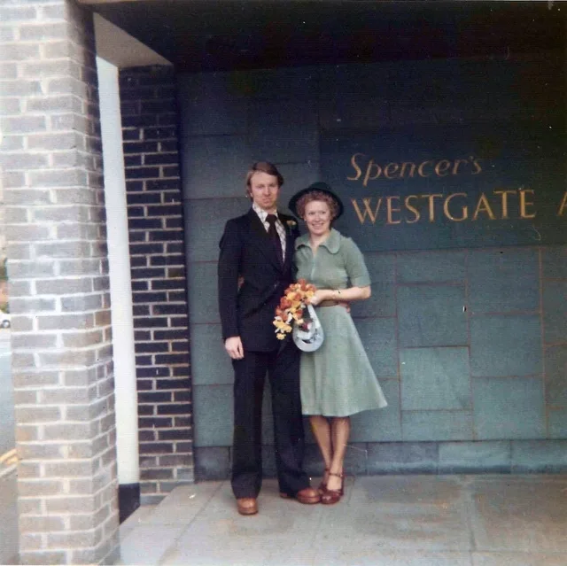 Iain and Pam on their wedding day, arm in arm outside the registry office, Iain in a dark suit and Pam in a green dress carrying a small posy.