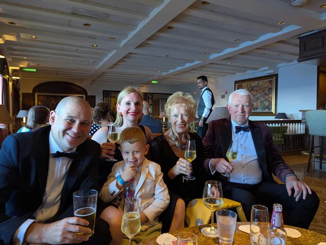Pam and Iain seated with family, all dressed smartly for a formal evening, raising glasses of champagne together.