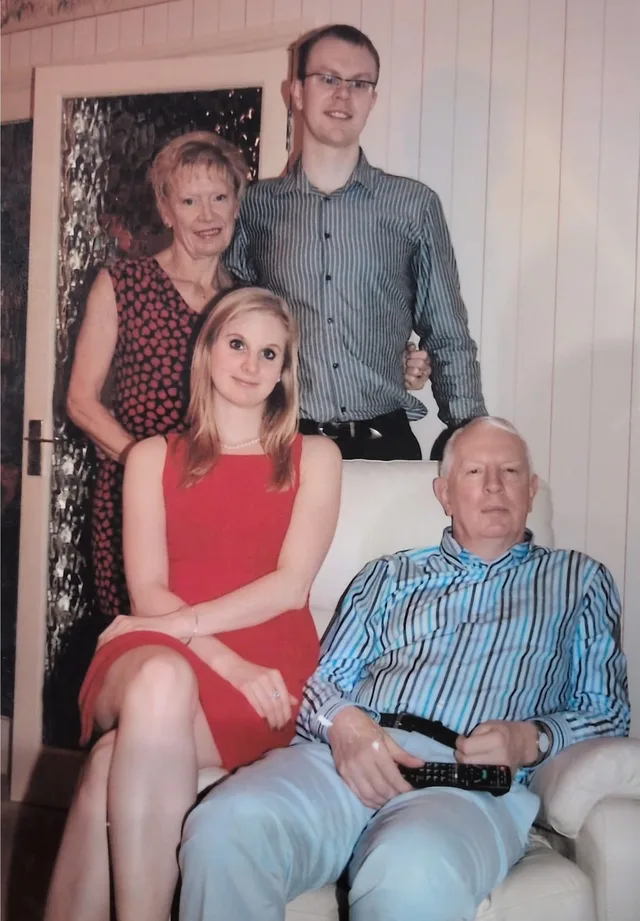 Pam and Euan standing behind the sofa with Anda seated in front, and Iain seated on the sofa holding the TV remote, at Christmas.